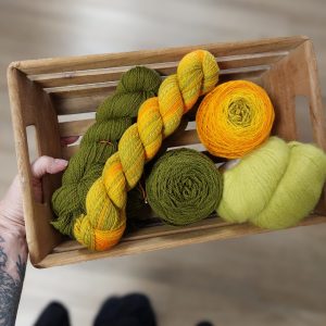 yarn in balls and skeins lays in a wooden crate. The yarns range in color from yellow to orange to green, in pale to dark shades 